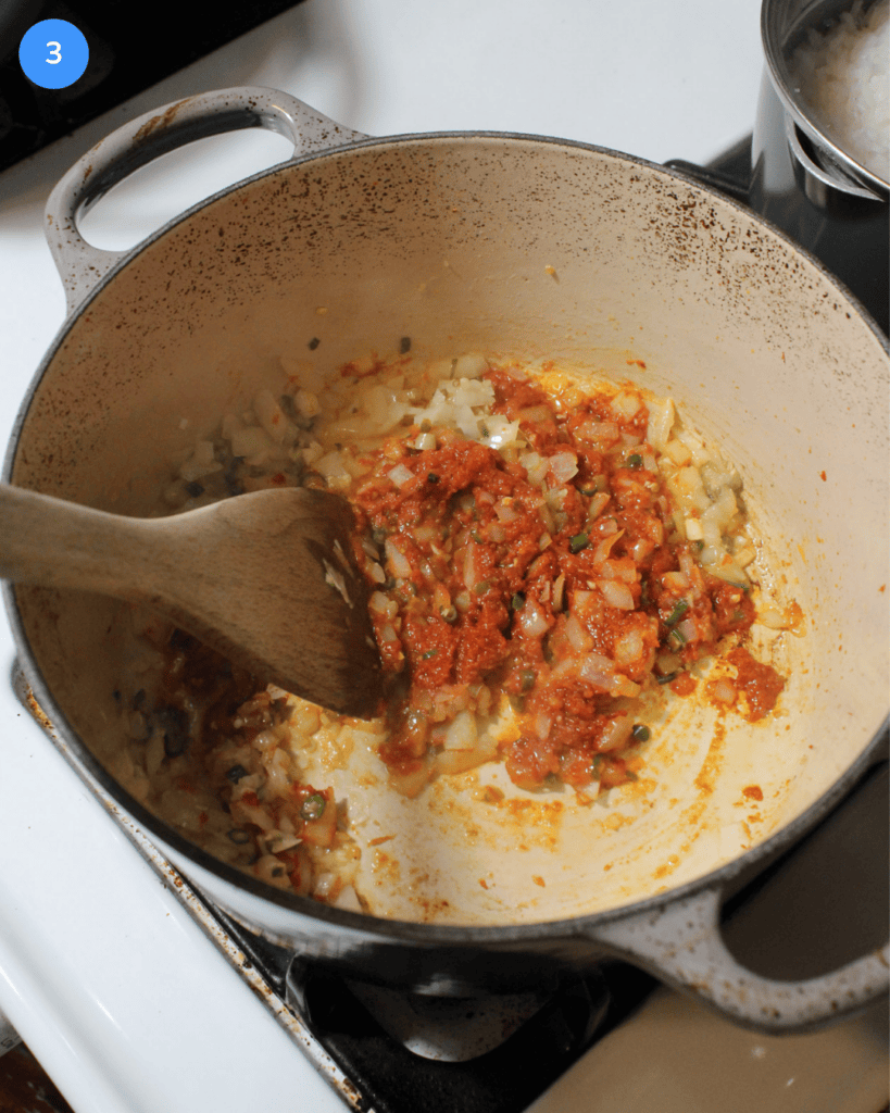 Onions, Thai chilies, garlic, ginger, and red curry paste being stirred together in a ceramic dutch oven on the stove.