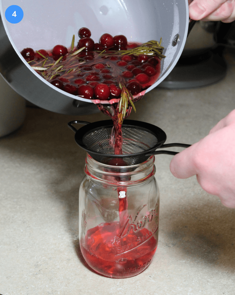 Pouring cranberry rosemary simple syrup from a pot into a mason jar, through a fine mesh sieve.