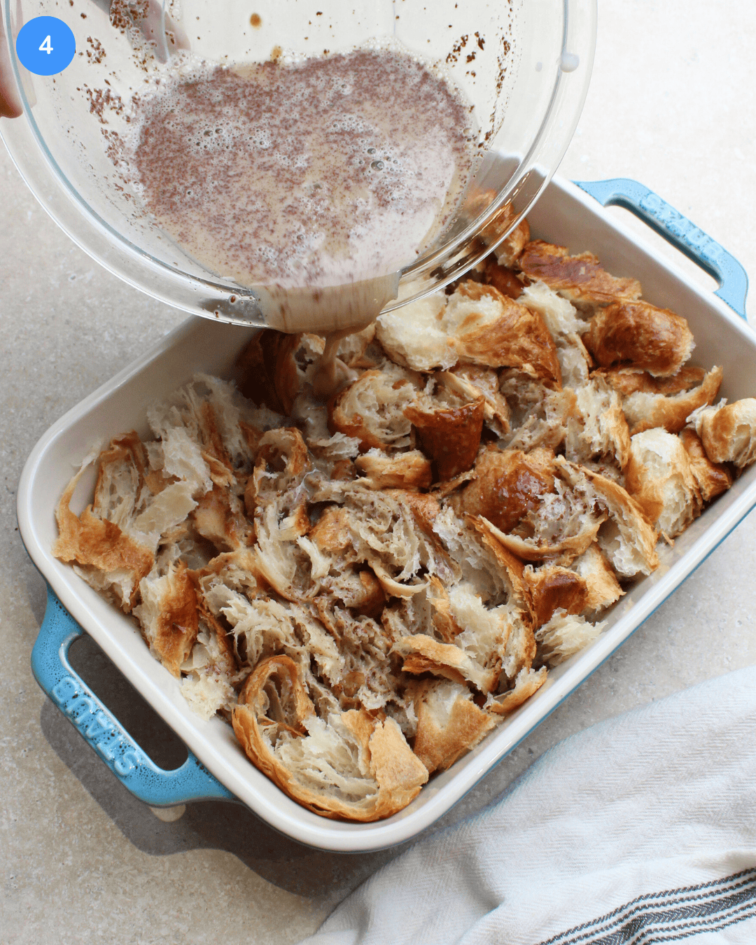 Ripped up croissant pieces in a blue casserole dish with french toast custard being poured over it.