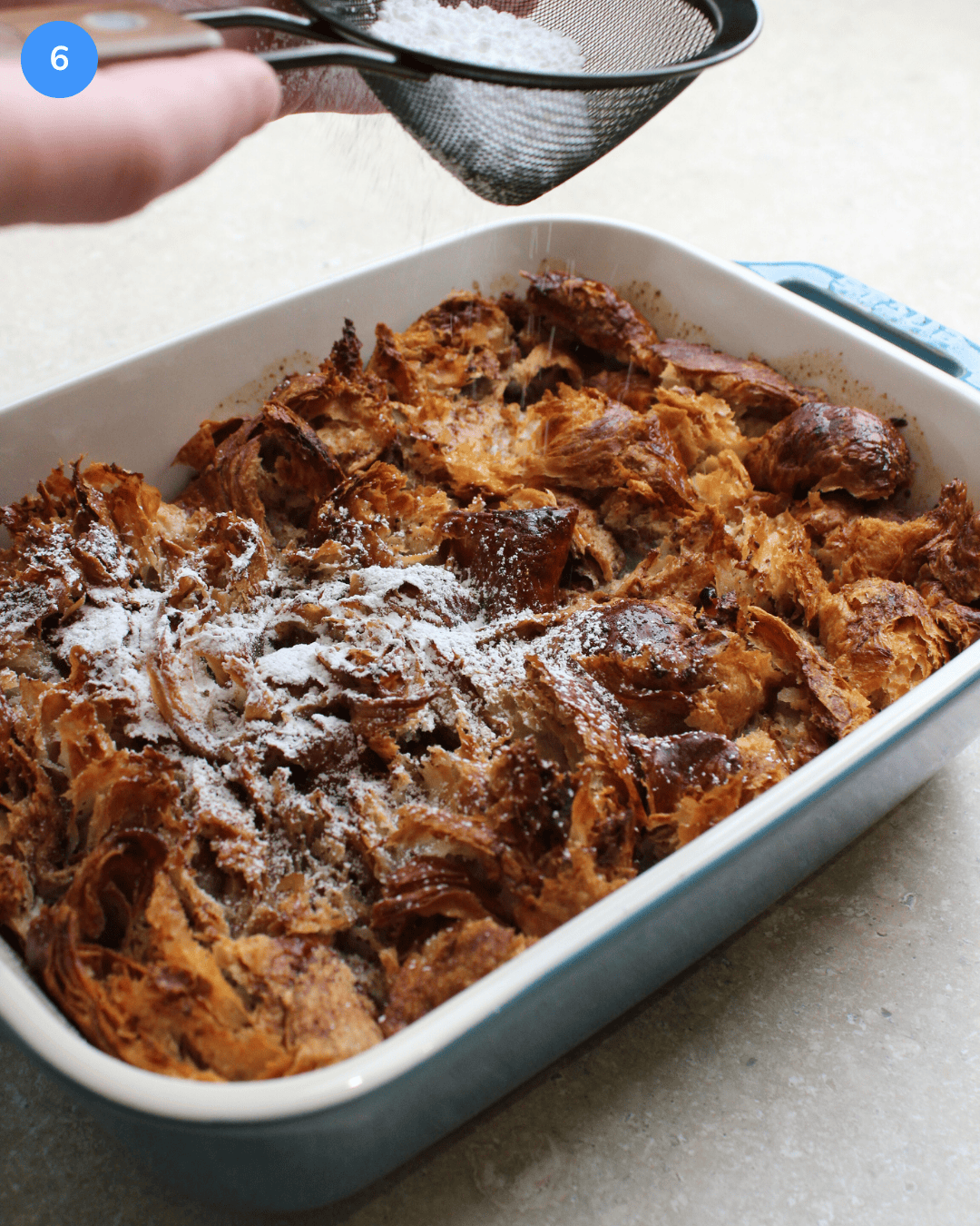 A freshly baked casserole dish of croissant french toast with powdered sugar being dusted over the top through a fine mesh sieve.