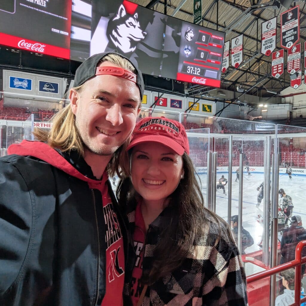 Nicole and Alex of The Candid Cooks at a Northeastern University hockey game, standing in front of the ice rink.