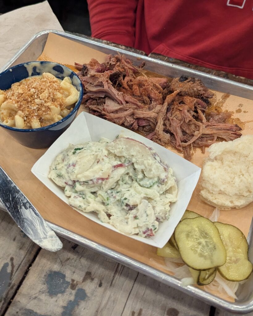 A pulled pork platter with potato salad, pickles, and mac n cheese on a silver tray.