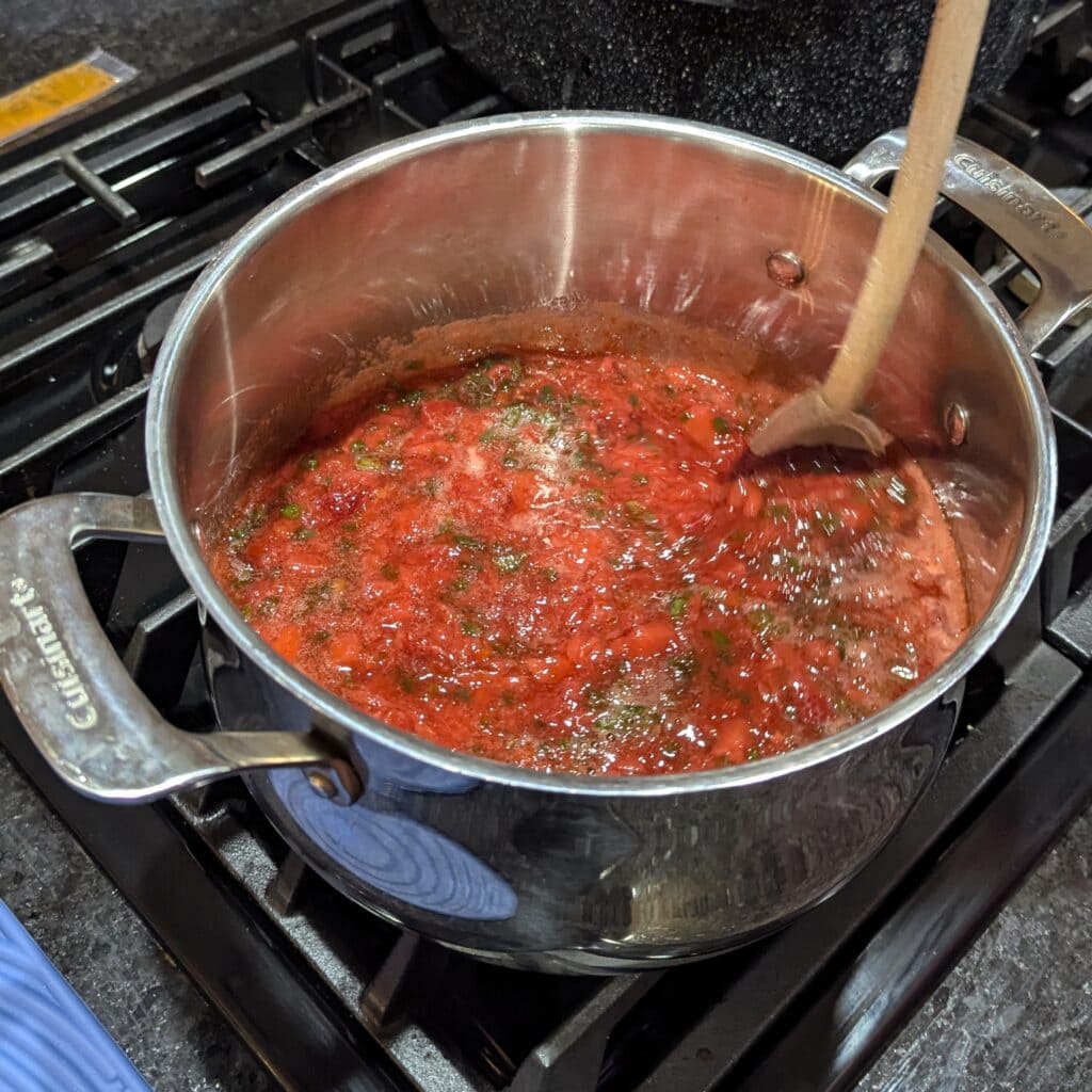 Strawberry hot pepper jelly cooking in a silver pot on the stove.
