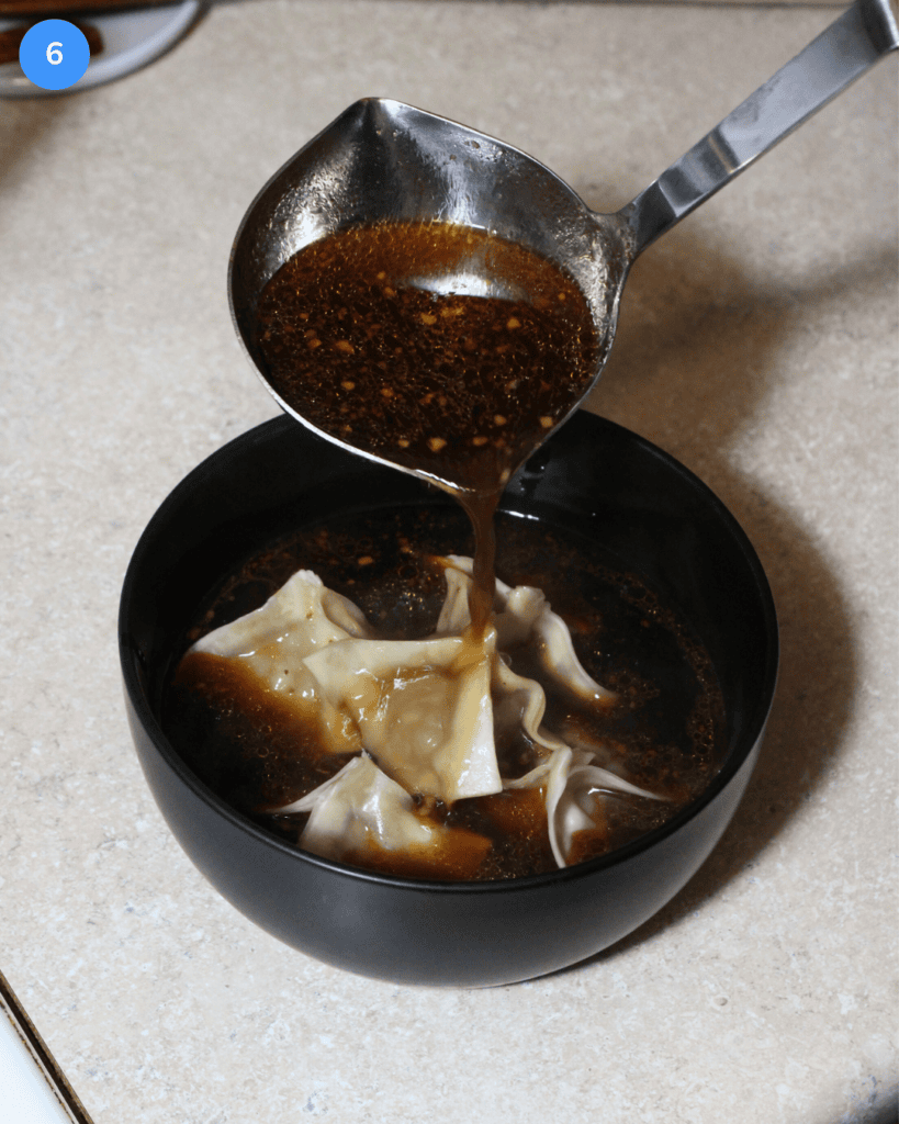 A ladle of Szechuan soup going into a black bowl with pork dumplings.