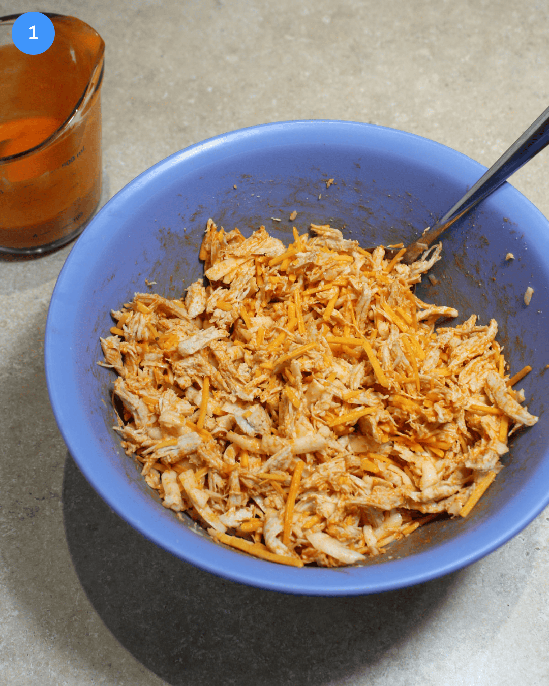A blue bowl filled with a mixture of shredded chicken, buffalo sauce, shredded cheddar, and shredded mozzarella.