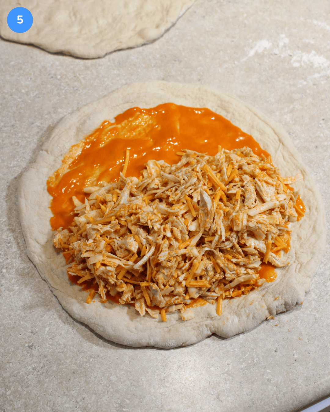 Unbaked pizza dough with buffalo sauce, shredded chicken, and cheese on it.