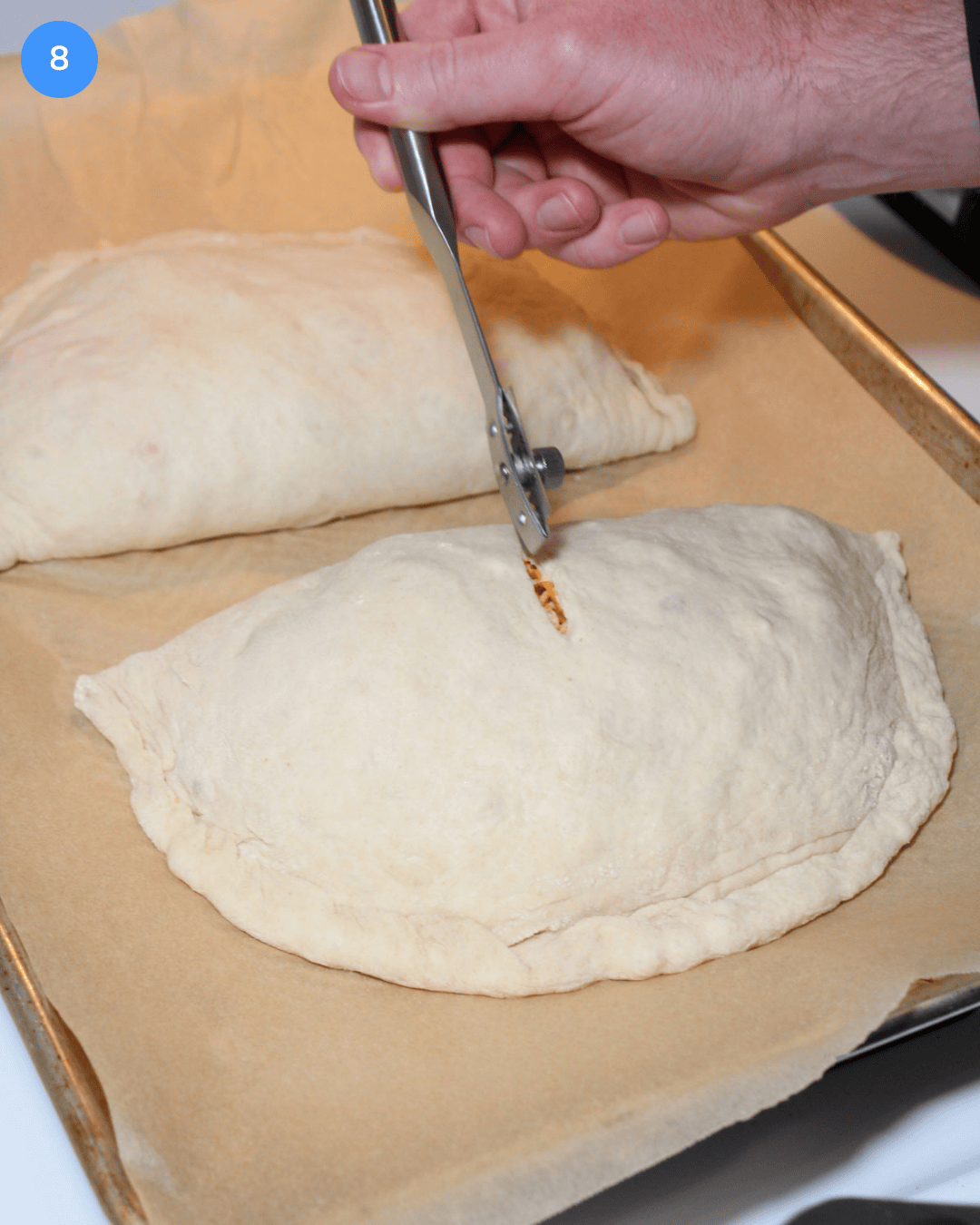 An unbaked calzone being vented with a bread lame on a parchment lined baking sheet before going into the oven.
