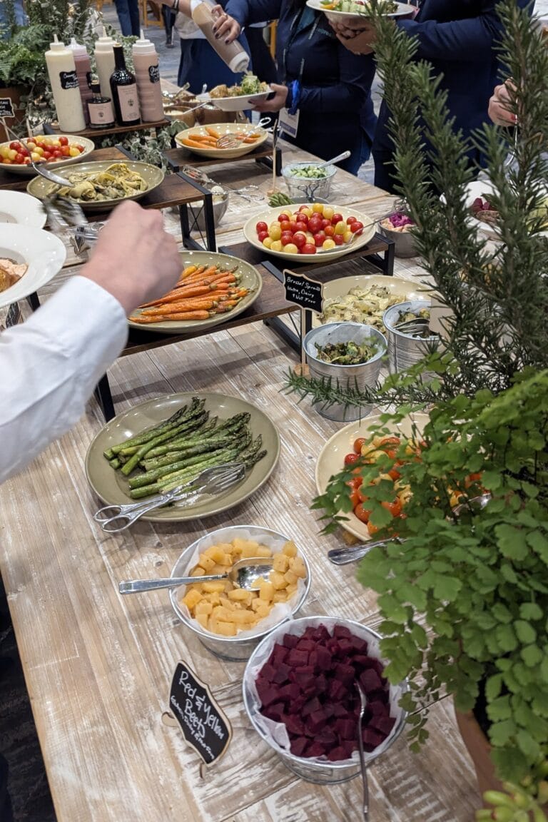 A colorful lunch buffet with roasted carrots, asparagus, tomatoes, and other vegetables on a light wood table.