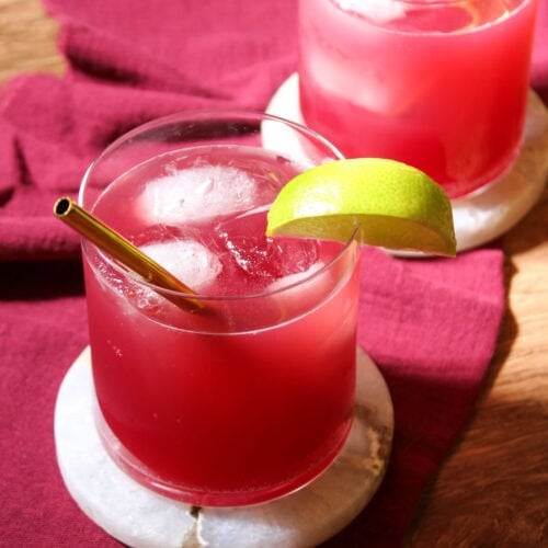 Two rocks glasses filled with pomegranate mocktails and ice, garnished with a lime wedge and a gold straw.