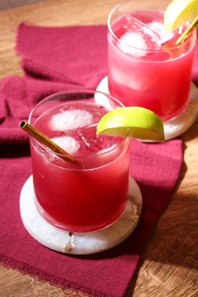 Two rocks glasses filled with pomegranate mocktails and ice, garnished with a lime wedge and a gold straw.