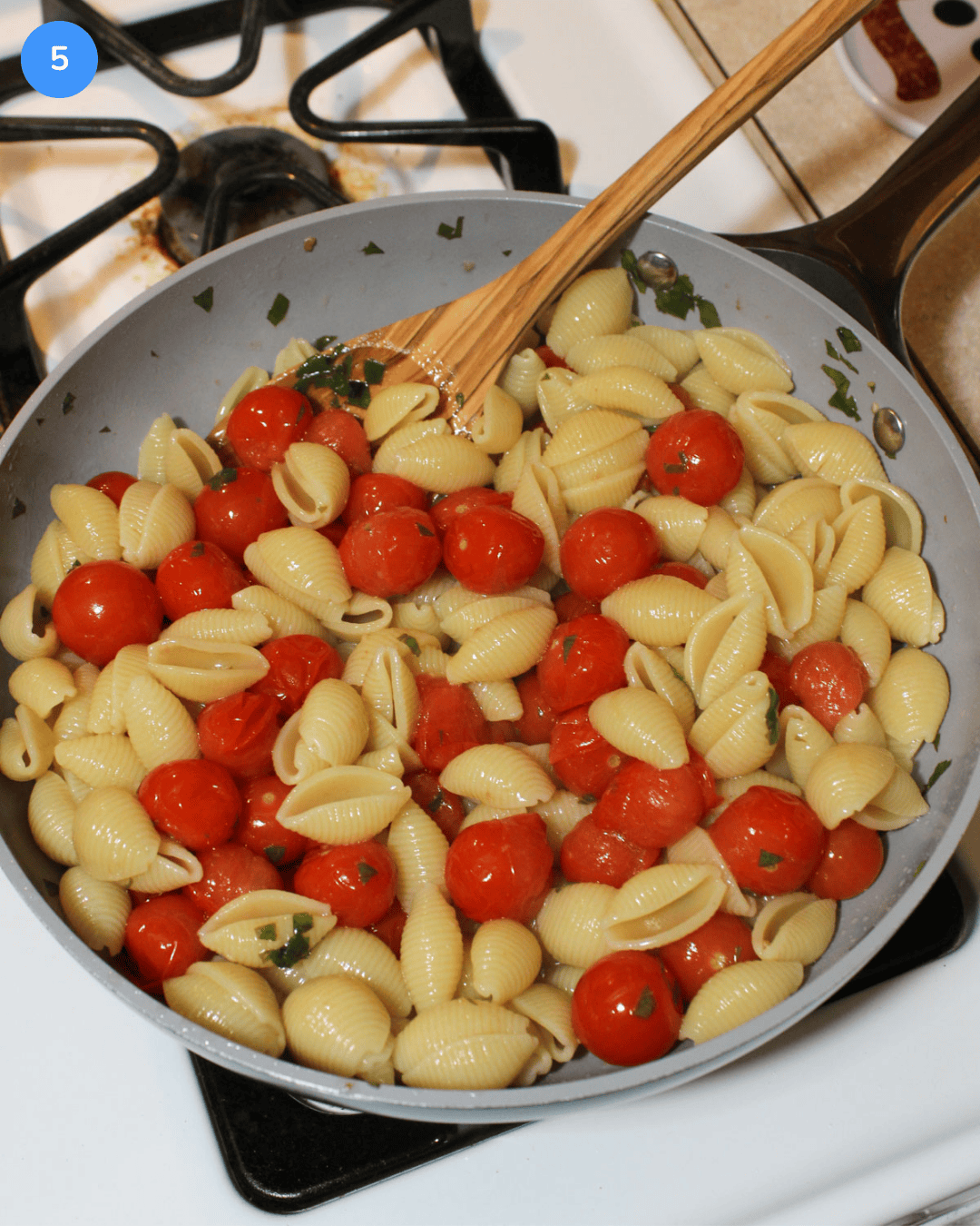 Pasta shells, blistered cherry tomatoes, and fresh basil in a nonstick ceramic frying pan on the stove with a wooden spoon.