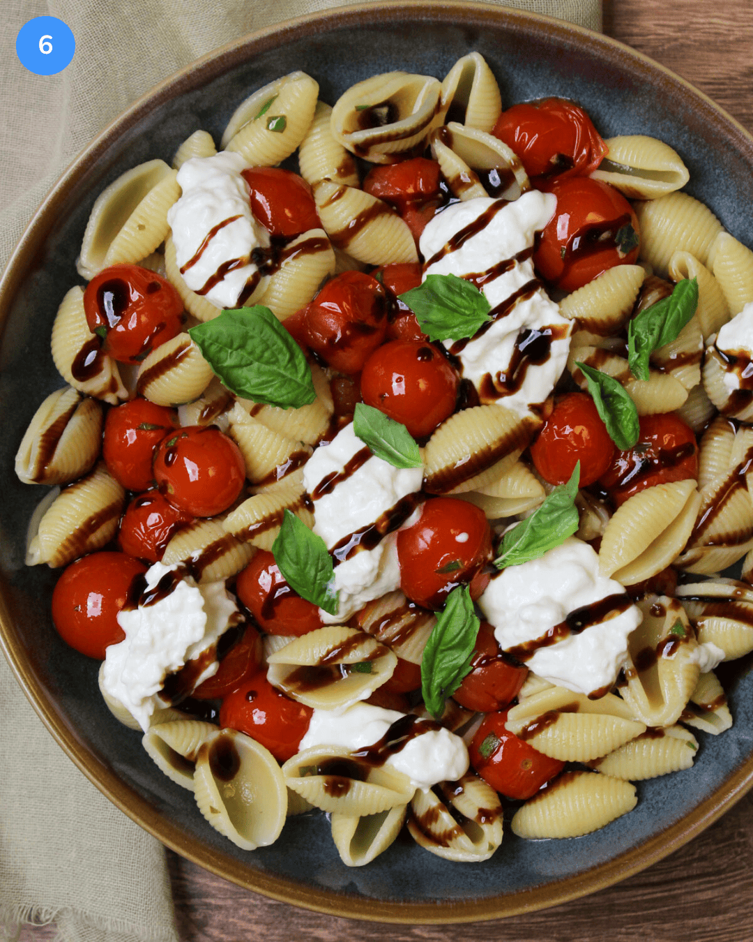 A bowl of pasta shells plated with blistered cherry tomatoes, dollops of burrata cheese, fresh basil leaves, and a drizzle of balsamic glaze.