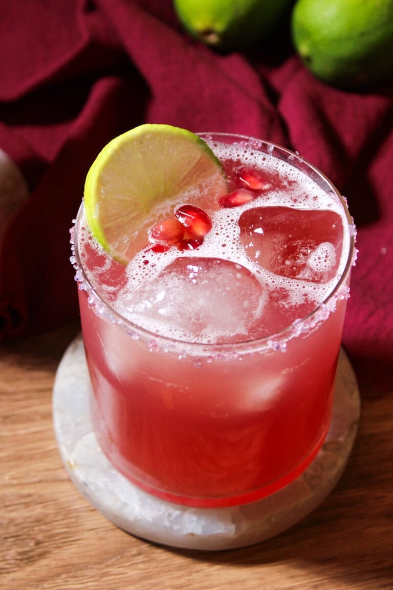 A freshly shaken pomegranate margarita on the rocks with a salted rim, lime wheel, and pomegranate arils for garnish.