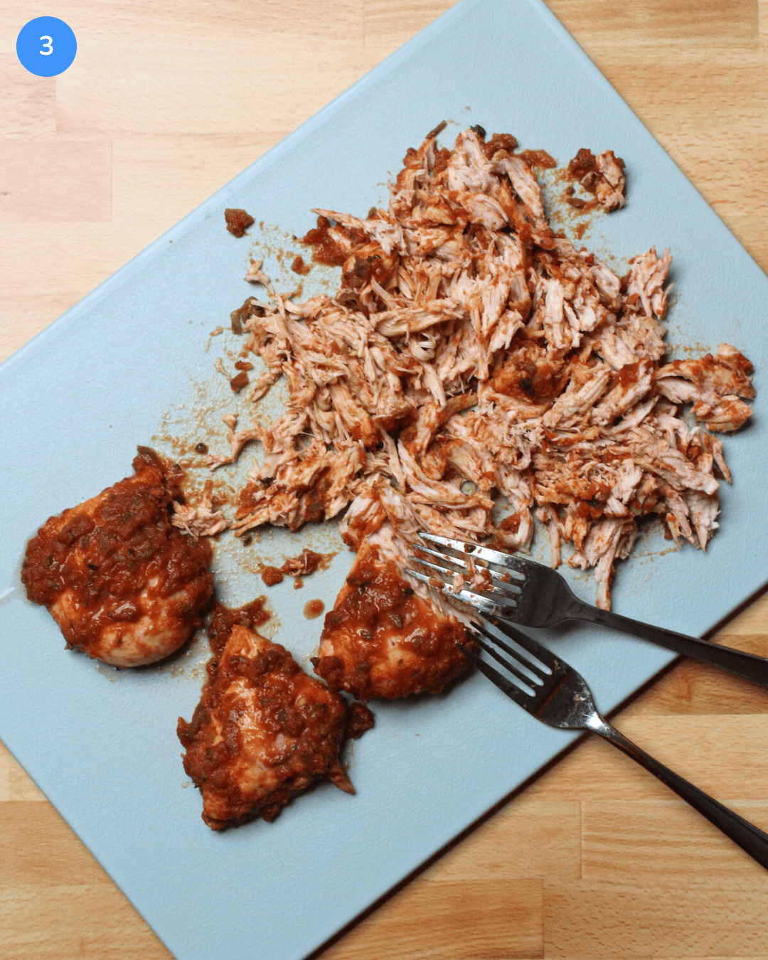 Freshly baked salsa marinated chicken breast being shredded with two forks on a cutting board.