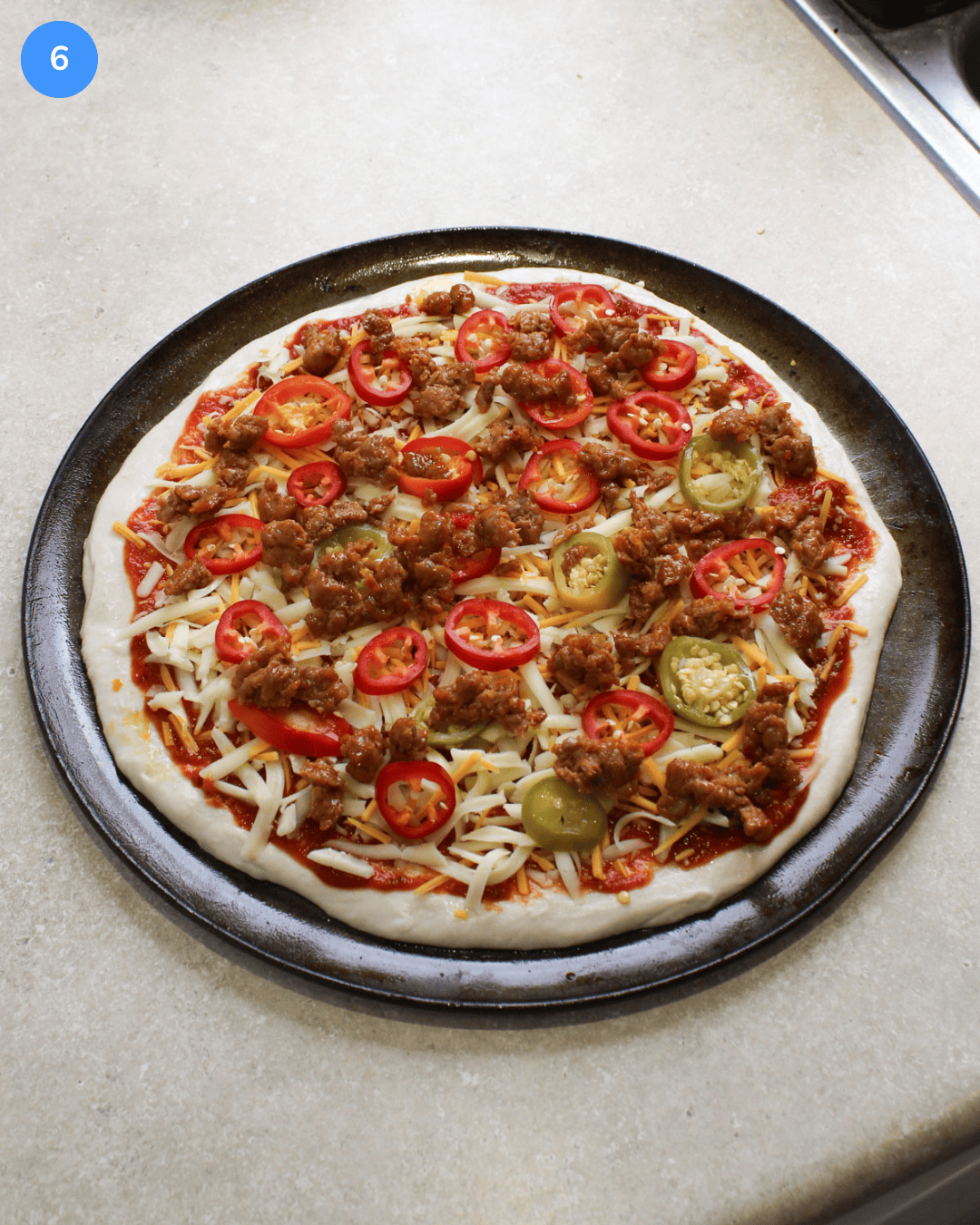 An uncooked pizza topped with spicy red sauce, shredded cheese, hot pepper slices, and ground Italian spicy sausage.