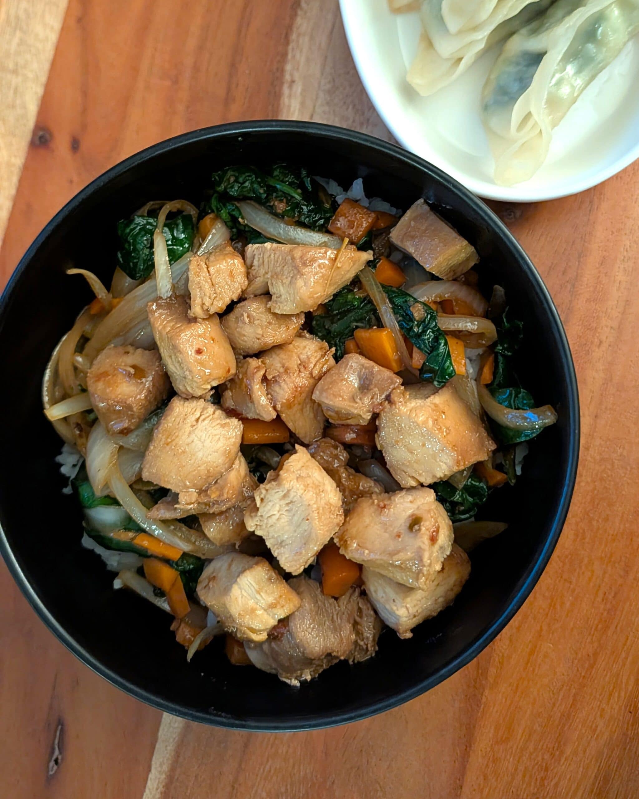 A chicken and veggie stir fry with rice in a black stoneware bowl, served with veggie dumplings on the side.