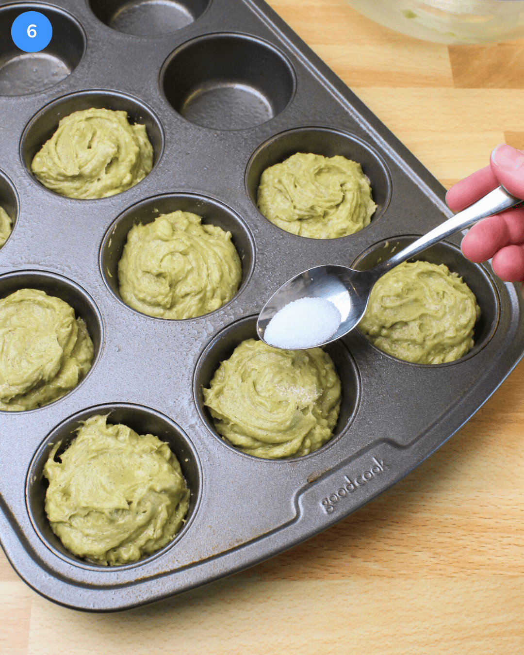 A spoonful of sugar being sprinkled on the tops of cinnamon avocado muffins in a muffin tin, before baking.