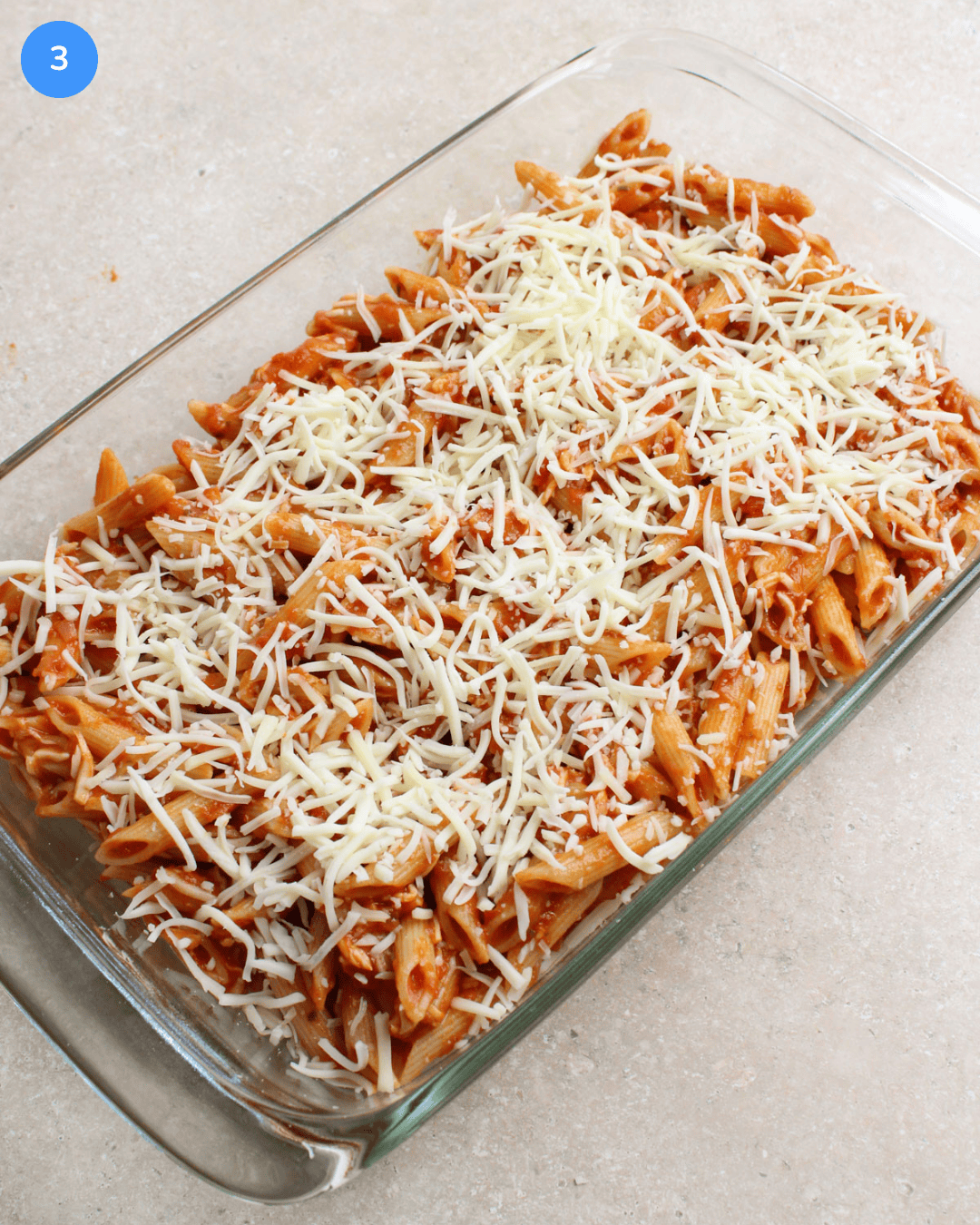 A 9 by 13 glass baking dish filled with penne pasta, red sauce, and shredded mozzarella cheese.