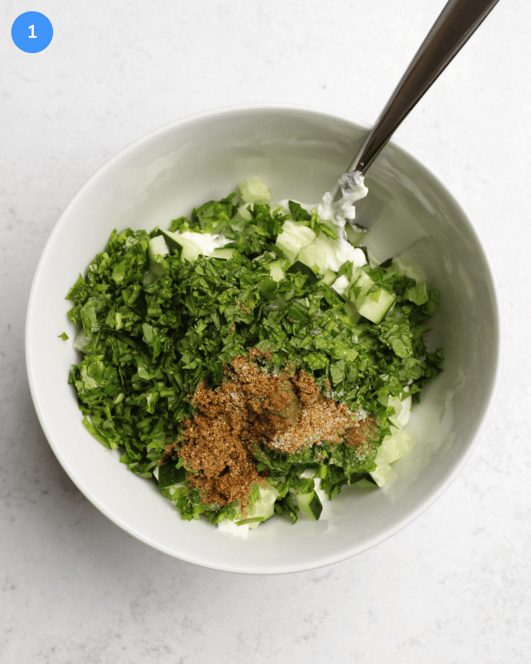 A white ceramic bowl with yogurt, fresh herbs, garam masala, salt, and diced cucumber.