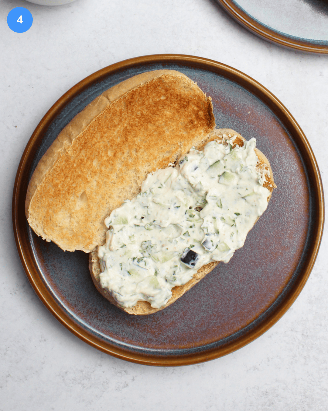 A freshly toasted sub roll with homemade cucumber raita slathered on the bottom half, on a blue stone plate.