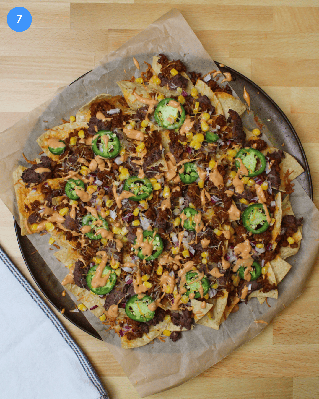 A round tray of freshly baked chorizo nachos topped with fresh veggies and a drizzle of chipotle mayo.