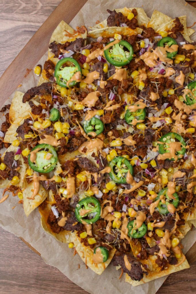 A wood serving tray lined with parchment paper of freshly baked chorizo nachos, topped with cheese, refried black beans, ground chorizo, fresh veggies, and a drizzle of chipotle mayo.
