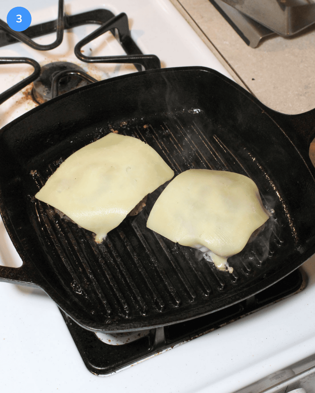 Two cheeseburger patties cooking on a cast iron grill pan on the stove.