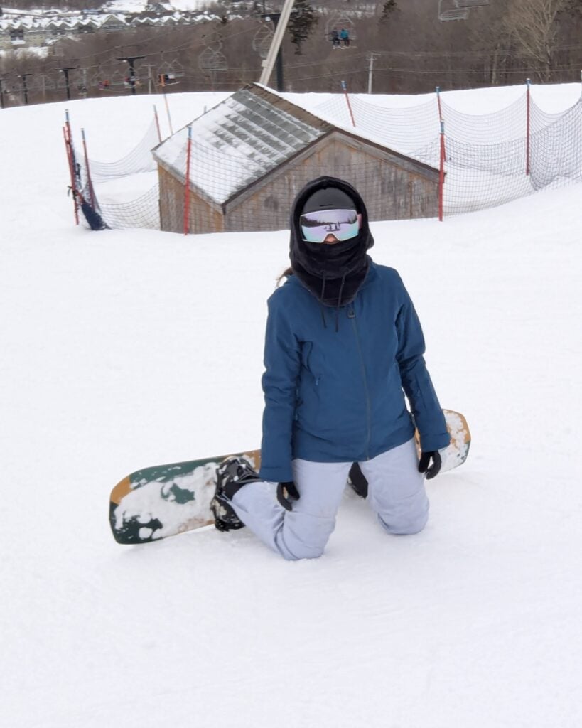 A photo of Nicole kneeling in the snow with her snowboard.
