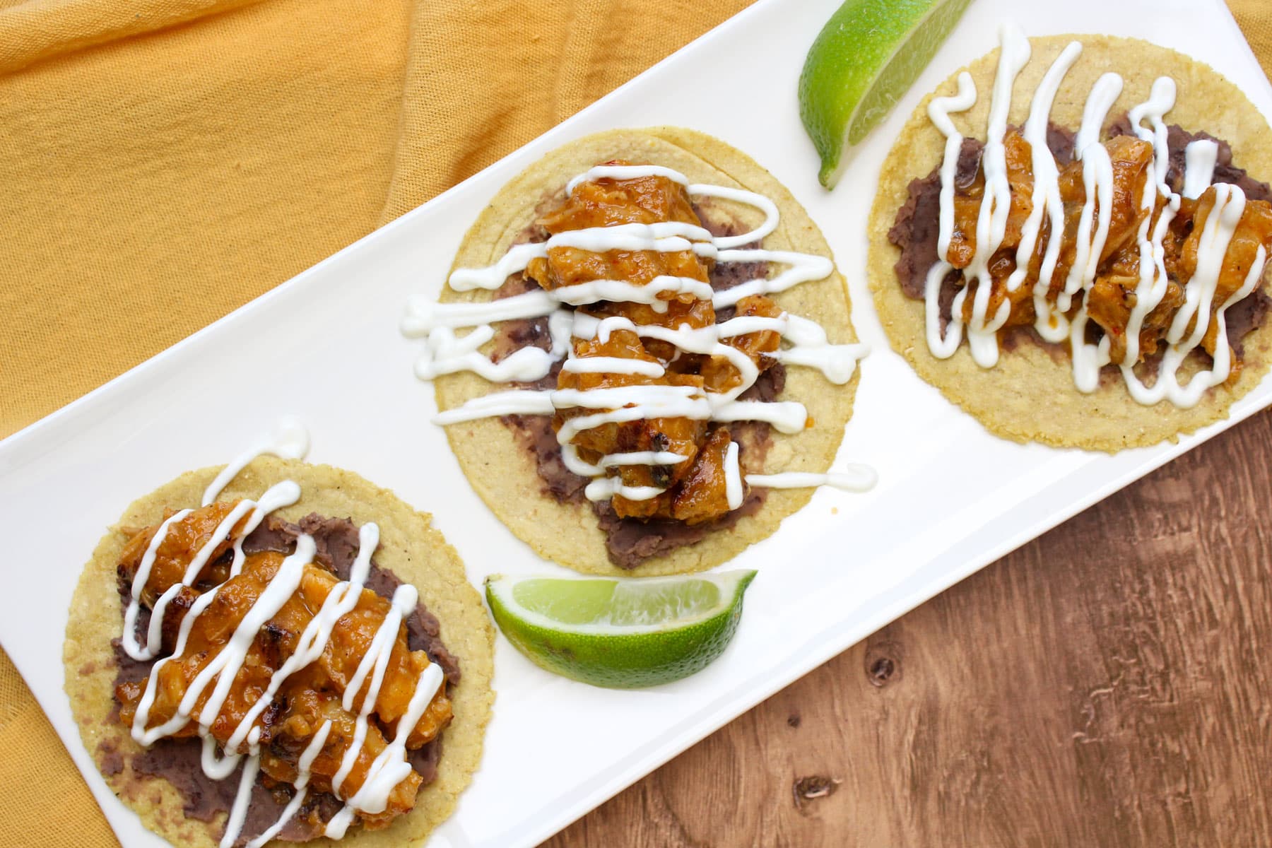 Three pineapple pork belly tacos on corn tortillas with refried black beans and lime crema drizzle on a white plate with lime wedges.