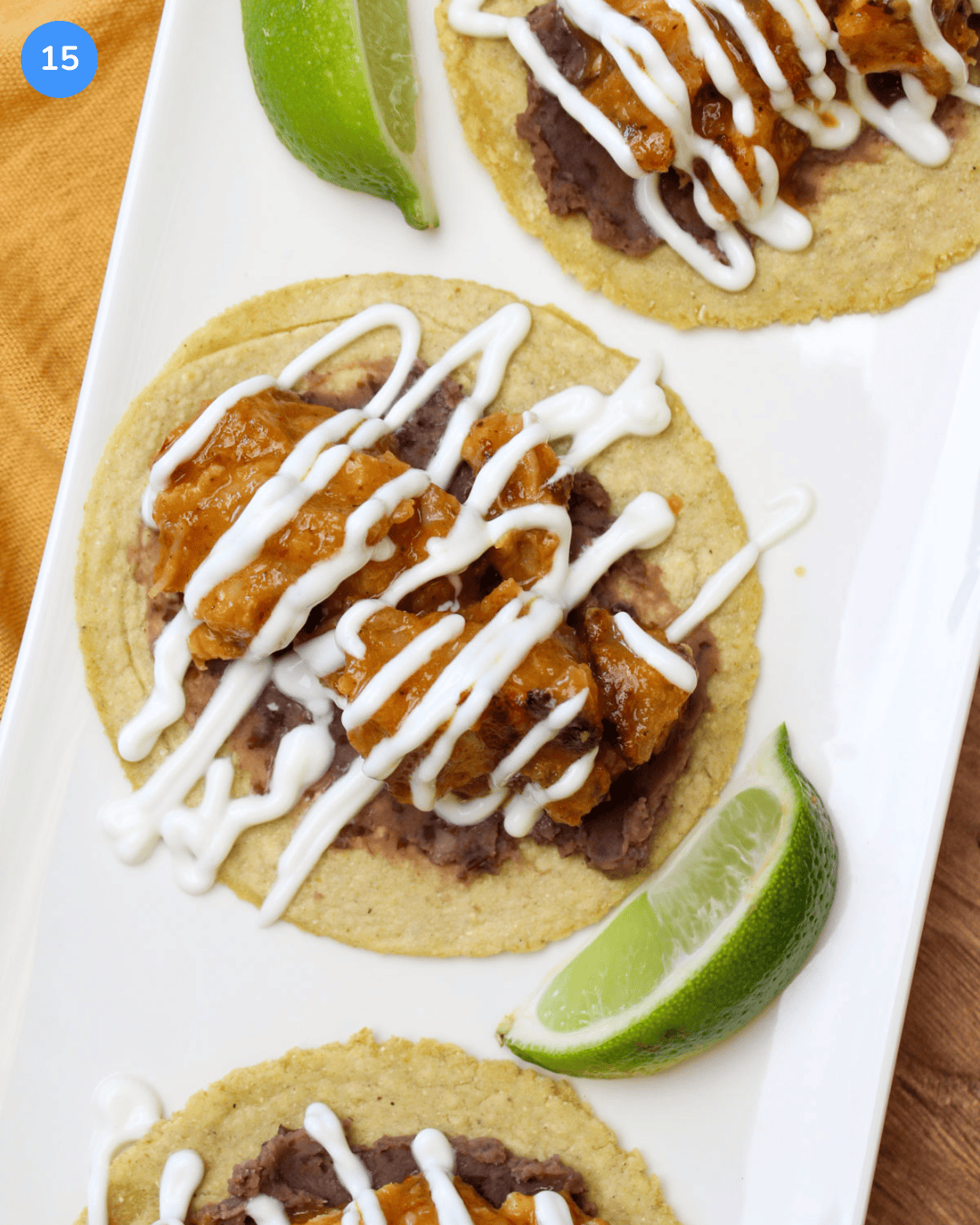 A crispy pork belly taco in pineapple chipotle sauce on a corn tortilla with refried black beans, lime crema drizzle, and a lime wedge.