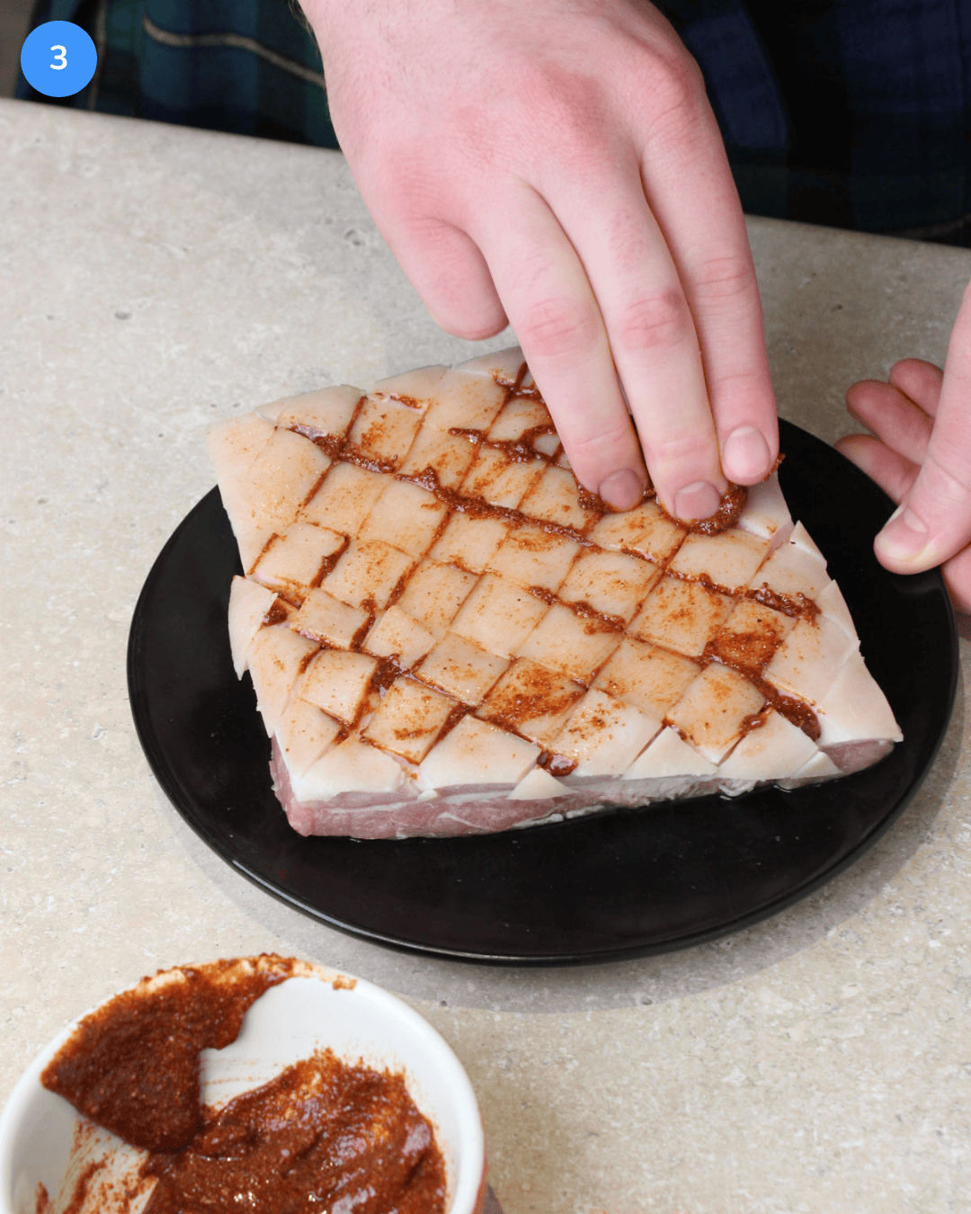 A slab of raw pork belly that's been scored through the skin in a cross-hatch pattern, being rubbed with a seasoned paste.