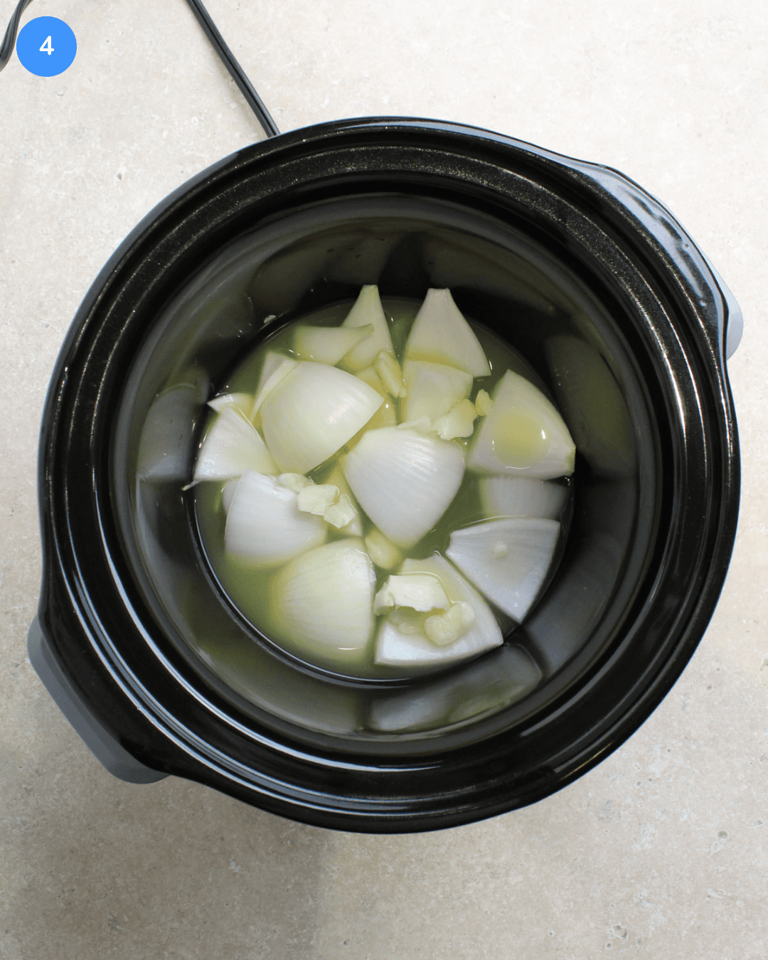 A black crockpot with white onion quarters, crushed garlic, lime juice, and pineapple juice on the bottom.