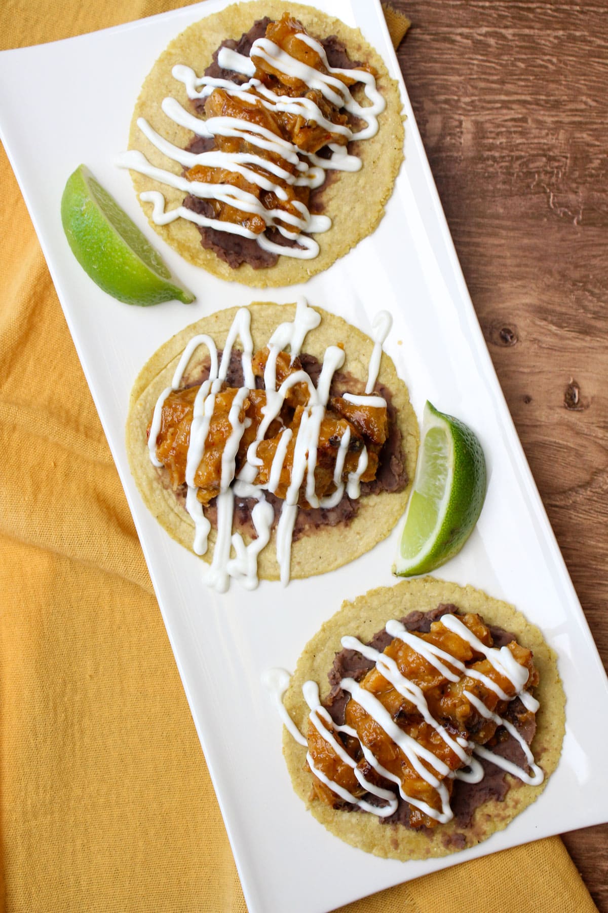 Freshly made crispy pork belly tacos with homemade corn tortillas, refried black beans, lime crema, and a pineapple chipotle sauce on a white plate with lime wedges.