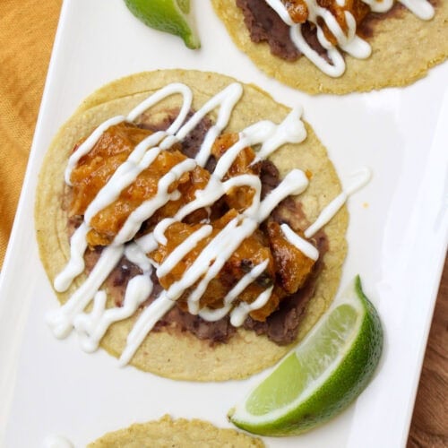 Crispy pork belly tacos on corn tortillas with refried black beans, lime crema drizzle, and lime wedges.