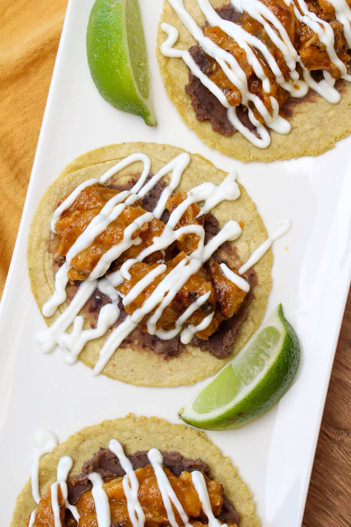 Crispy pork belly tacos on corn tortillas with refried black beans, lime crema drizzle, and lime wedges.
