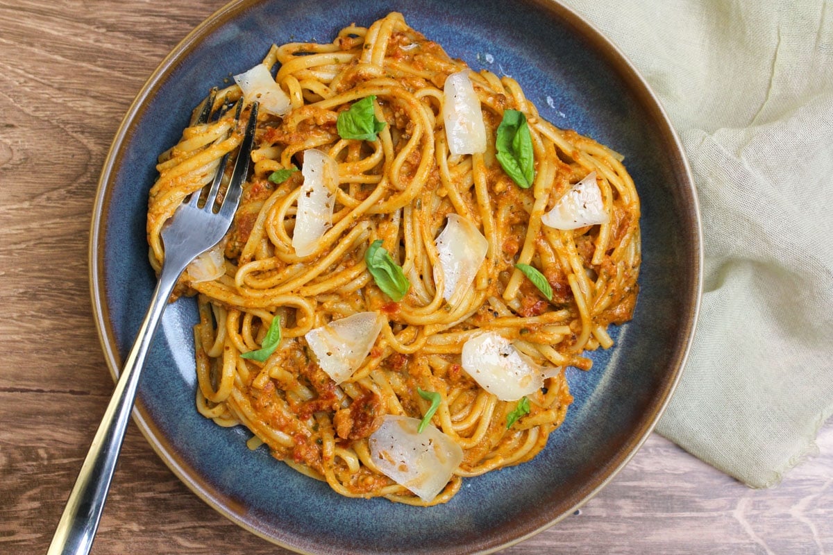 A blue stone bowl of sun dried tomato pesto linguine, topped with fresh basil leaves and parmesan shards.
