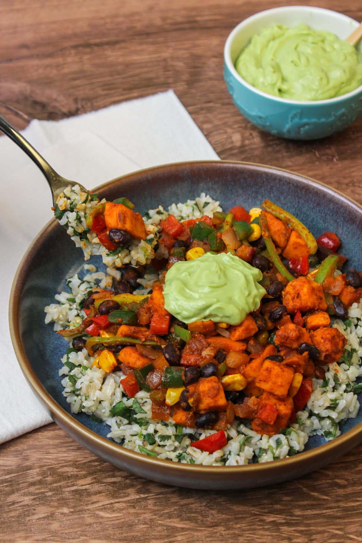 A forkful of sweet potato taco bowl being scooped out of a blue stone bowl with a small blue dish of avocado lime crema in the background.