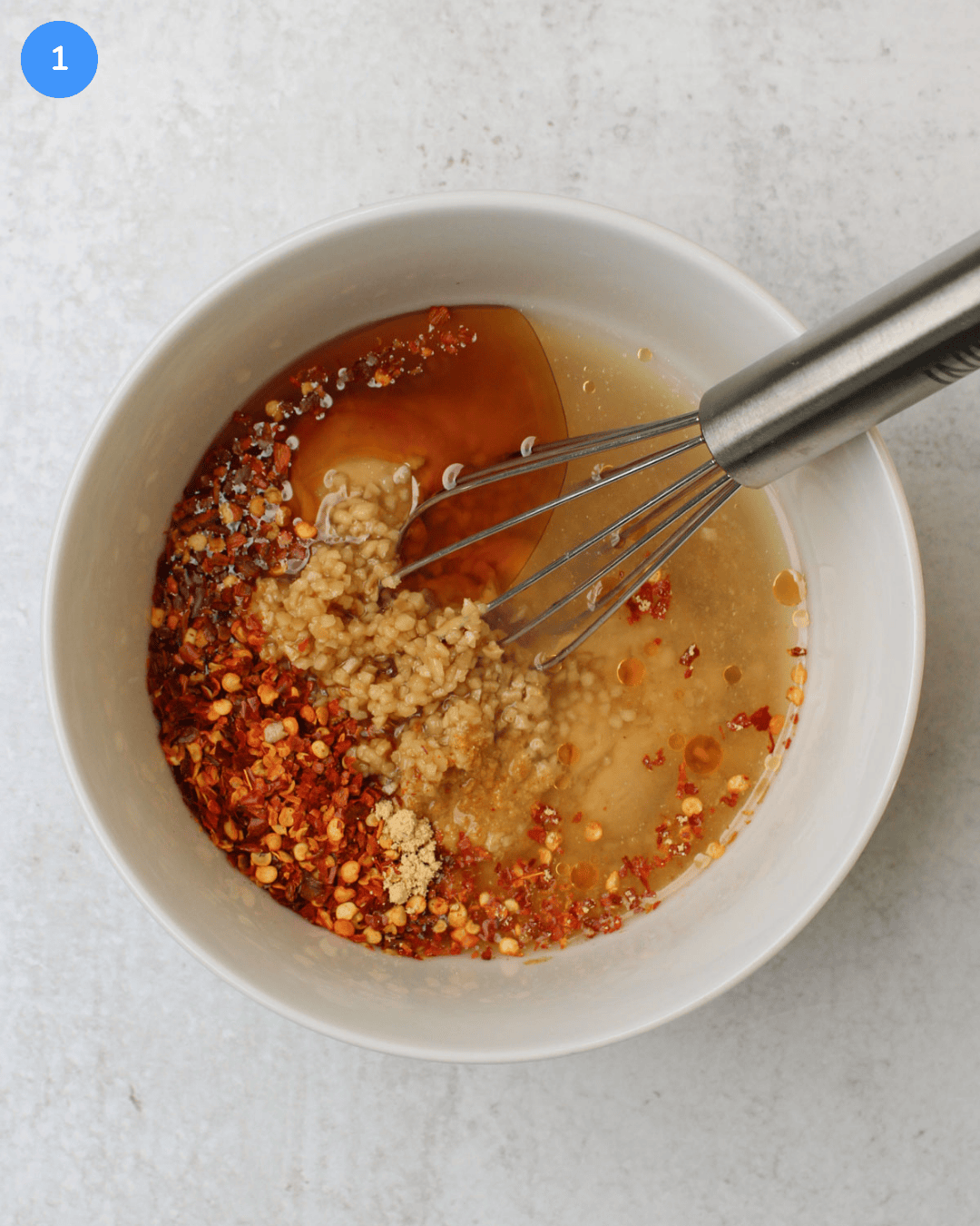 Cashew butter, maple syrup, sesame oil, rice vinegar, garlic, crushed red pepper, and other spices in a small white bowl with a mini whisk.