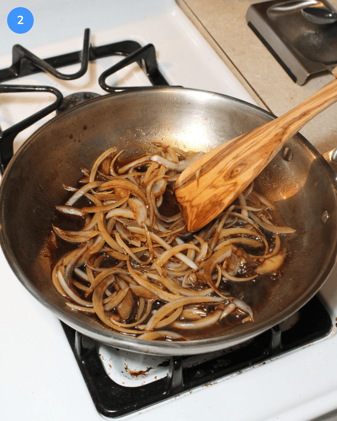 Sliced onions cooking in balsamic vinegar, salt, and sugar in a stainless steel frying pan on the stove.
