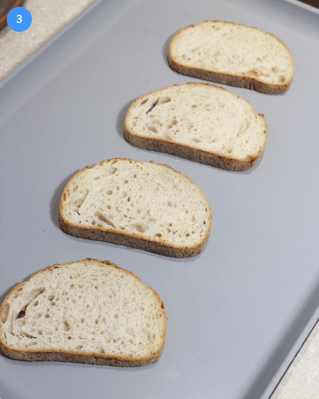 4 lightly toasted slices of Italian rustic bread on a ceramic baking sheet.