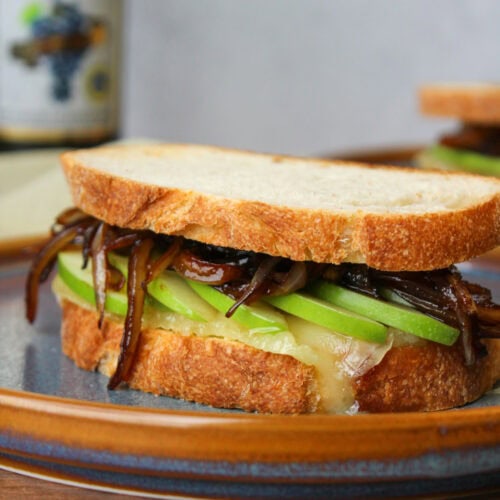 A gooey baked brie sandwich with green apple slices and balsamic onions on rustic Italian bread, served on a blue stone plate.