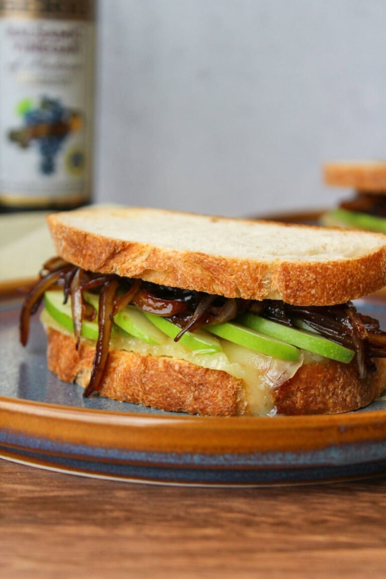 A gooey baked brie sandwich with green apple slices and balsamic onions on rustic Italian bread, served on a blue stone plate.