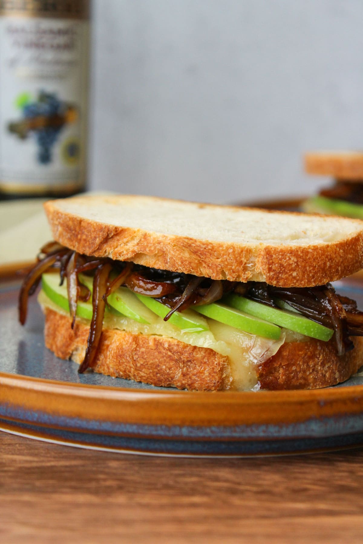 A gooey baked brie sandwich with green apple slices and balsamic onions on rustic Italian bread, served on a blue stone plate.