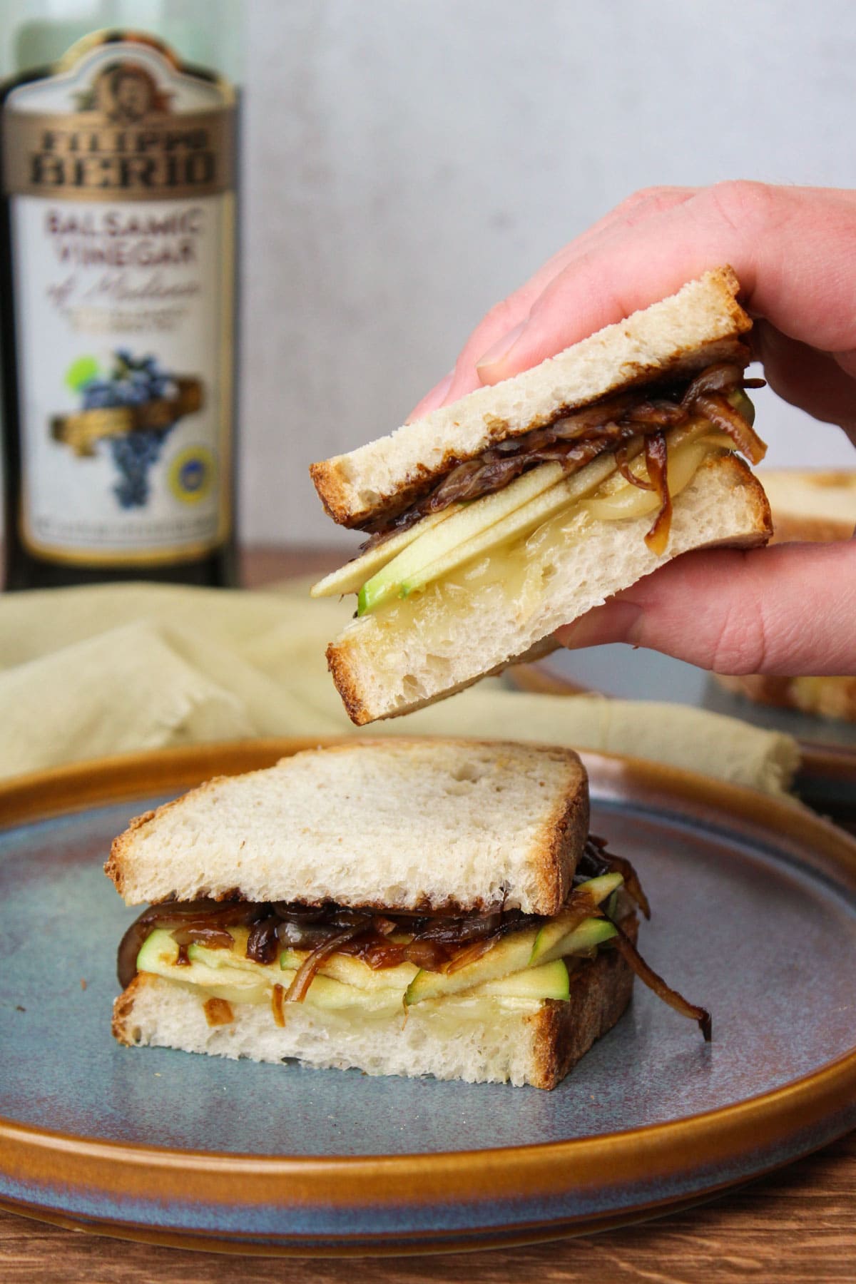 A hand picking up one half of an apple brie sandwich with balsamic onions from a blue stone plate.