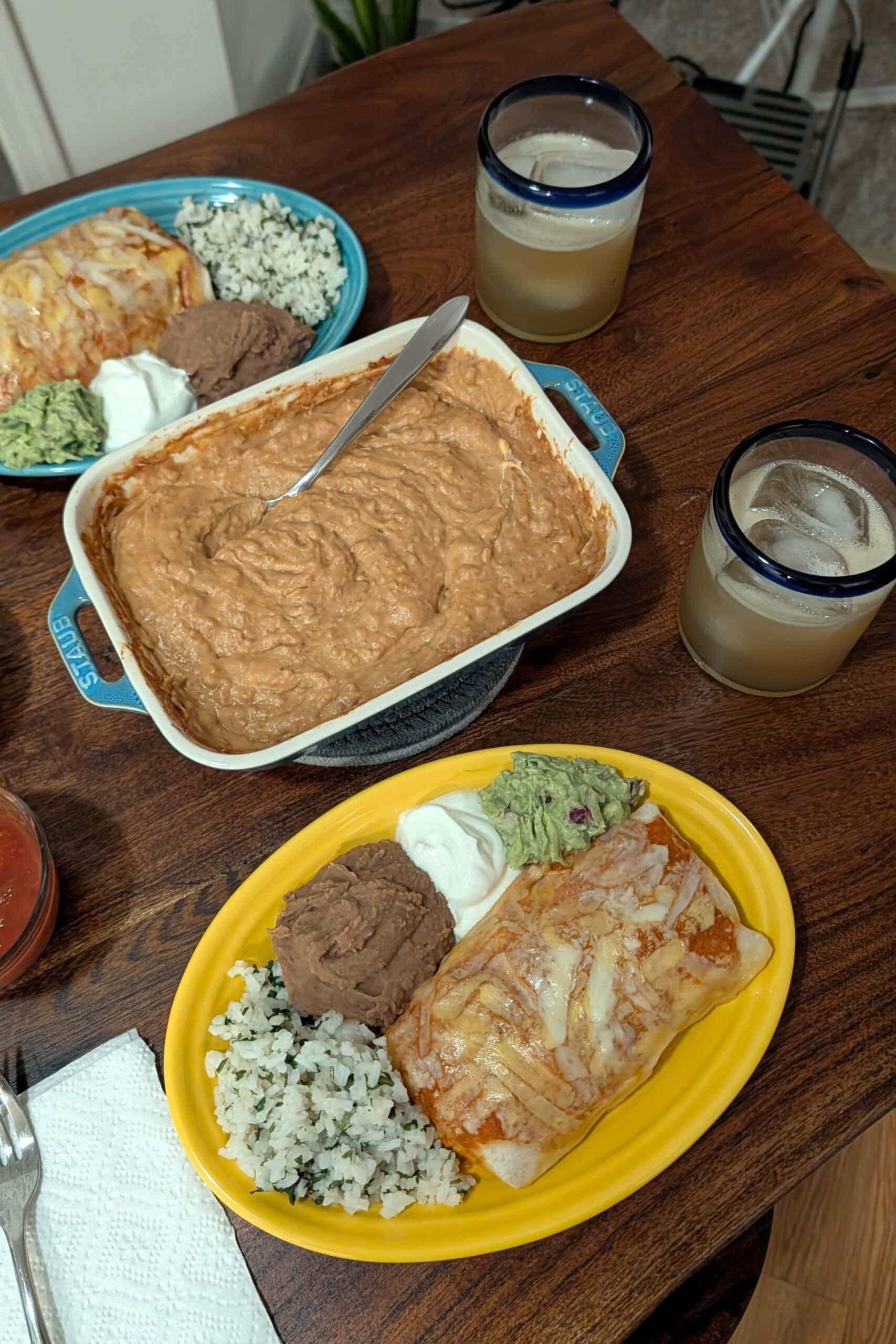 Two burrito plates and a baking dish of Mexican bean dip, and margaritas, on a wood dining table.