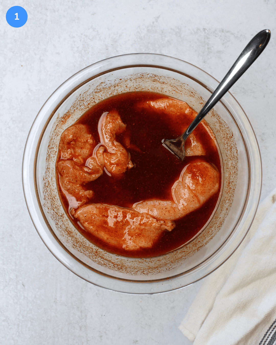 A glass mixing bowl of chicken breast marinating in lime juice, oil, salt, and Mexican seasonings.