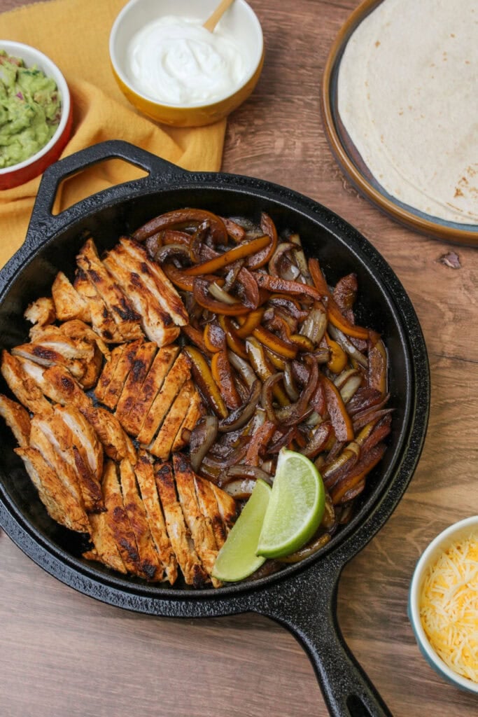 A cast iron skillet serving freshly made fajita chicken, alongside flour tortillas, greek yogurt, guacamole, and shredded cheese.