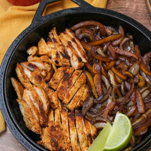 A cast iron skillet with freshly cooked fajita chicken, peppers and onions, and lime wedges, served with small bowls of guacamole and greek yogurt.