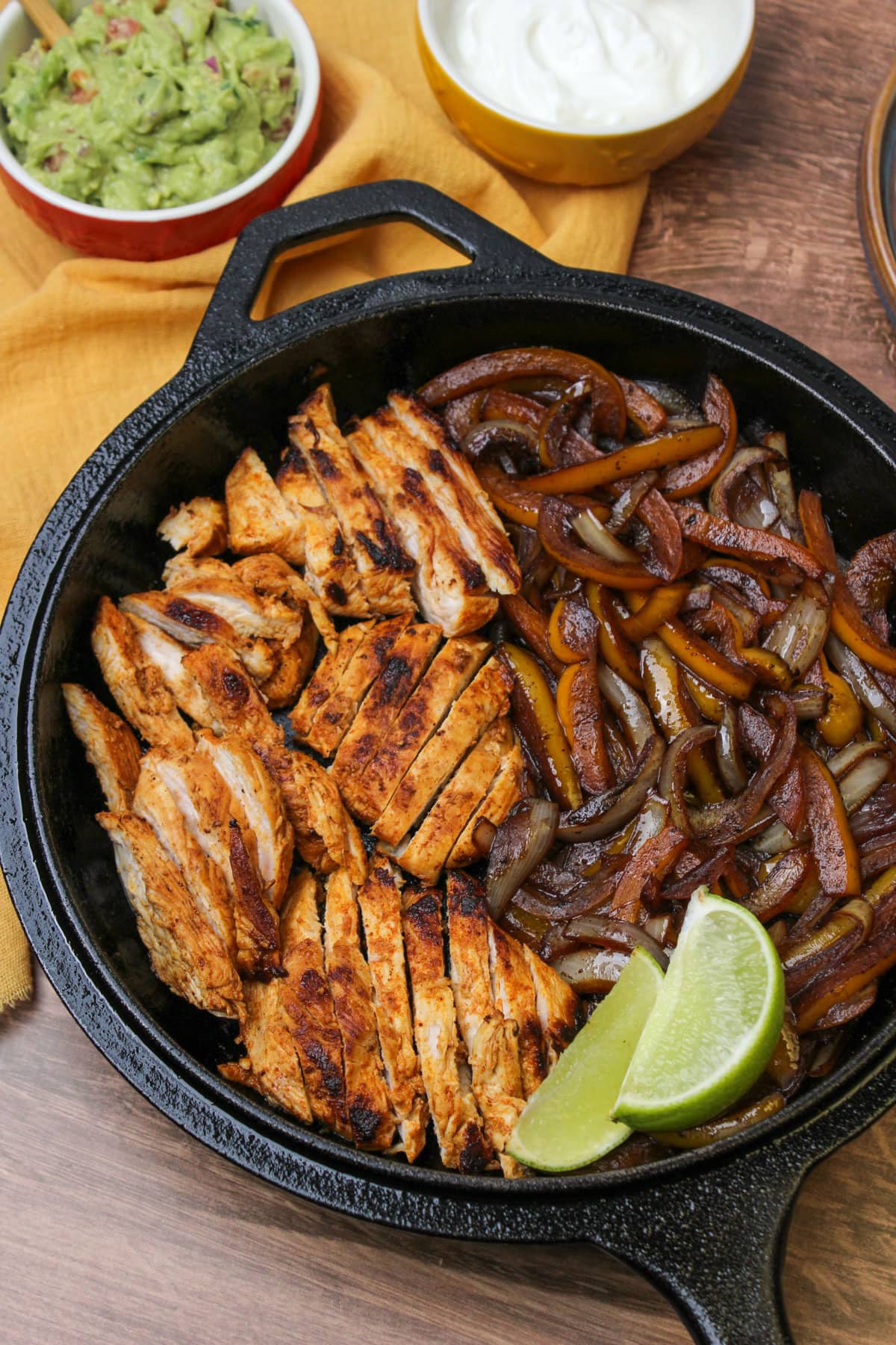 A cast iron skillet with freshly cooked fajita chicken, peppers and onions, and lime wedges, served with small bowls of guacamole and greek yogurt.