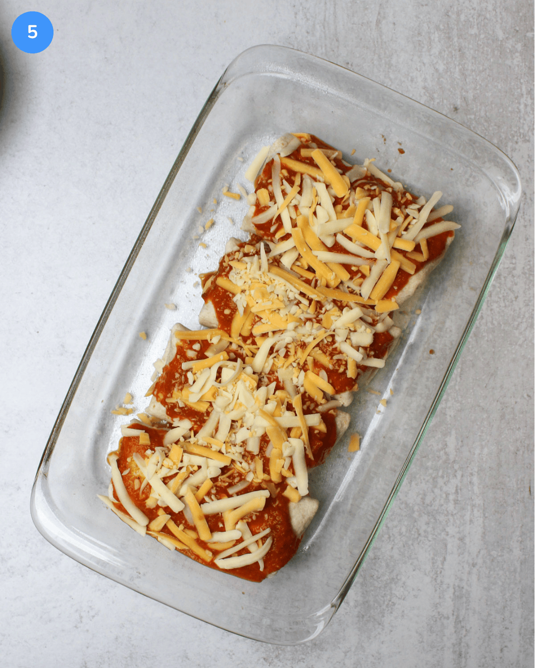 Four burritos covered with ranchero sauce and Mexican cheese in a glass baking dish.